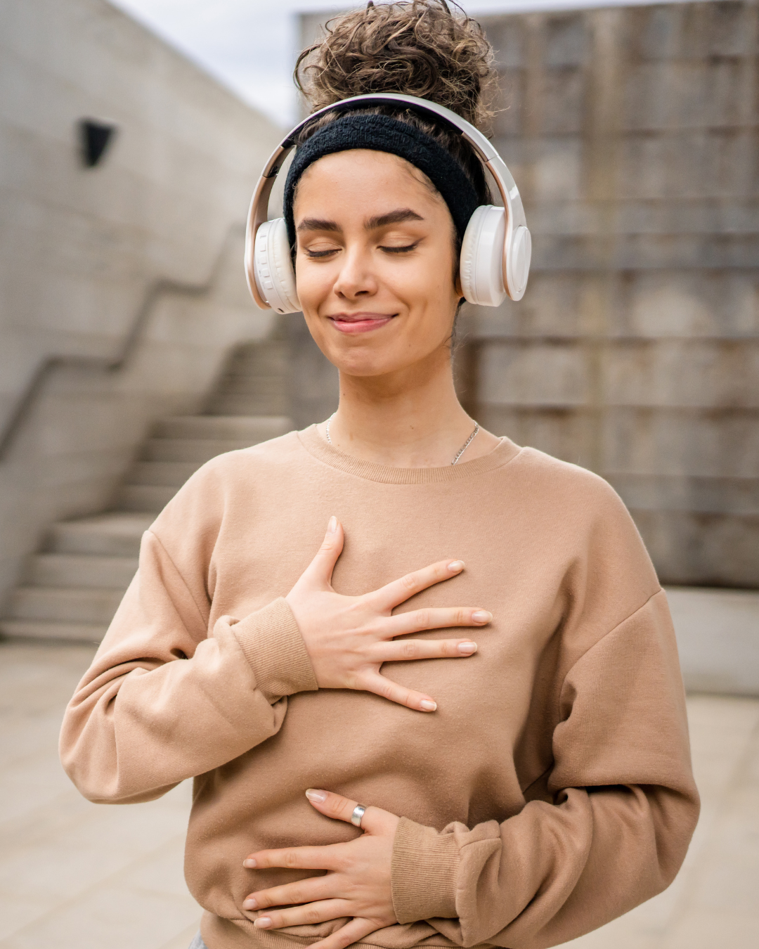 Woman wearing headphones and a beige sweatshirt, standing outdoors with a neutral background