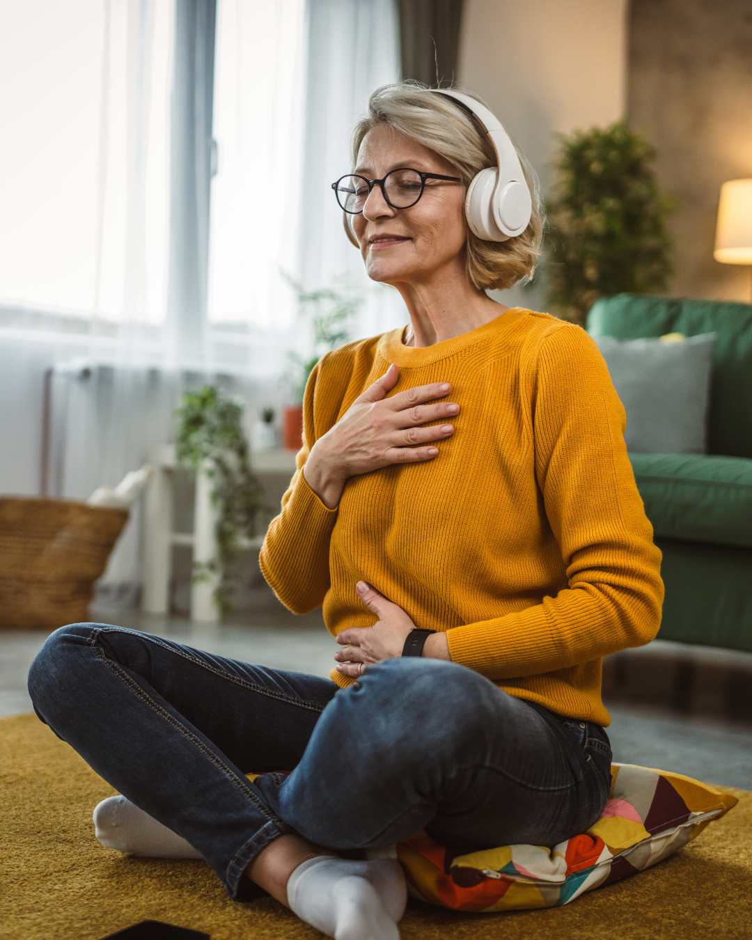 Woman wearing headphones and a yellow sweater sitting on a colorful rug in a living room.