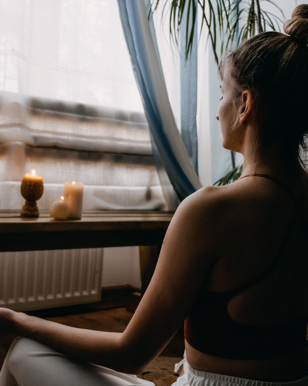Woman sitting by a window with candles and plants, creating a serene atmosphere.