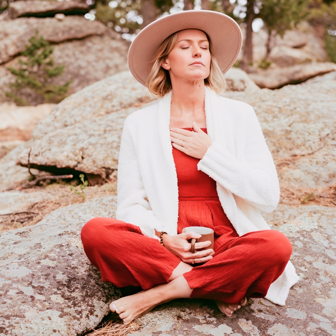 Woman in red outfit and beige hat sitting on a rock with eyes closed, holding a cup.