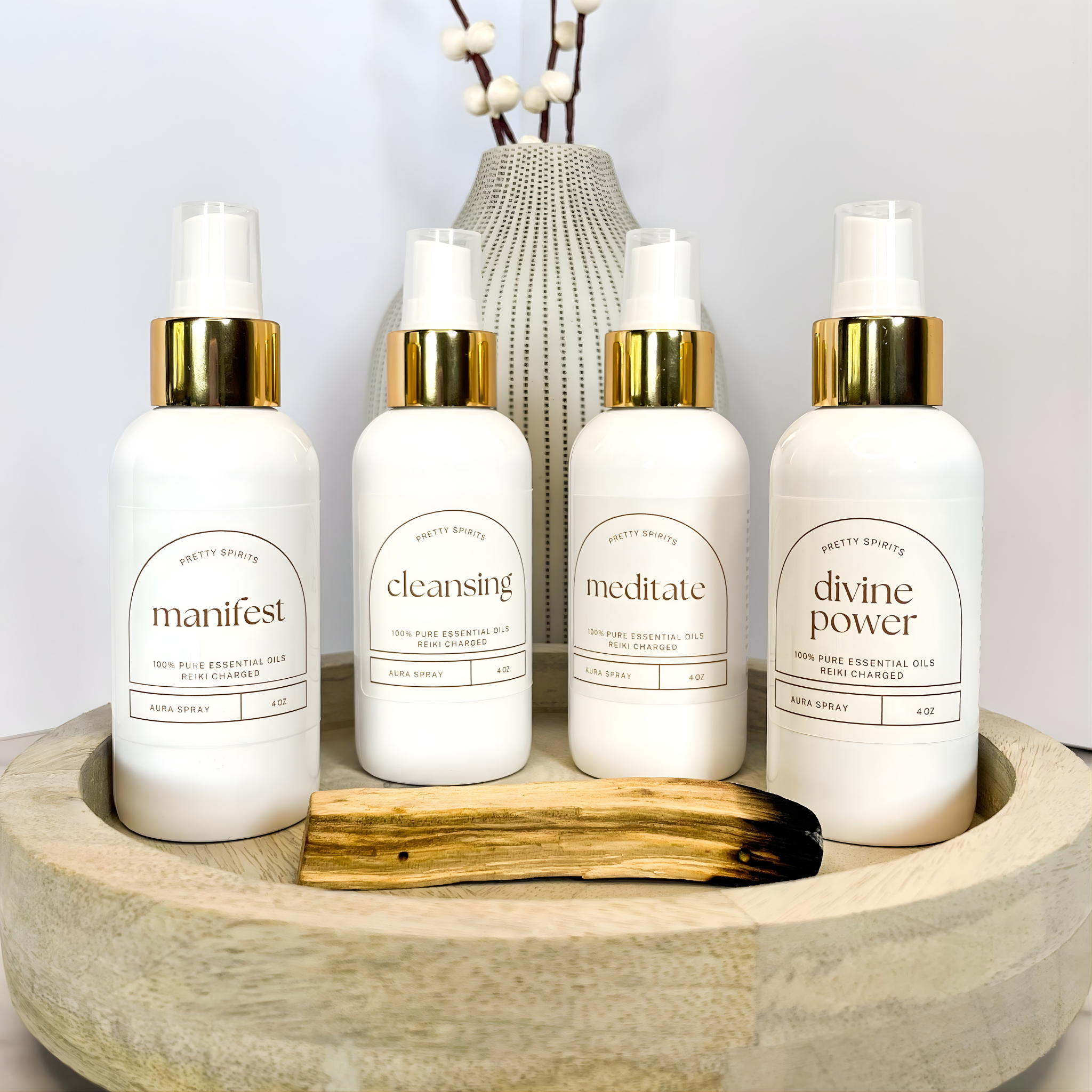 Four white bottles with gold caps on a wooden tray, featuring labels like 'manifest', 'cleansing', 'meditate', and 'divine power'.