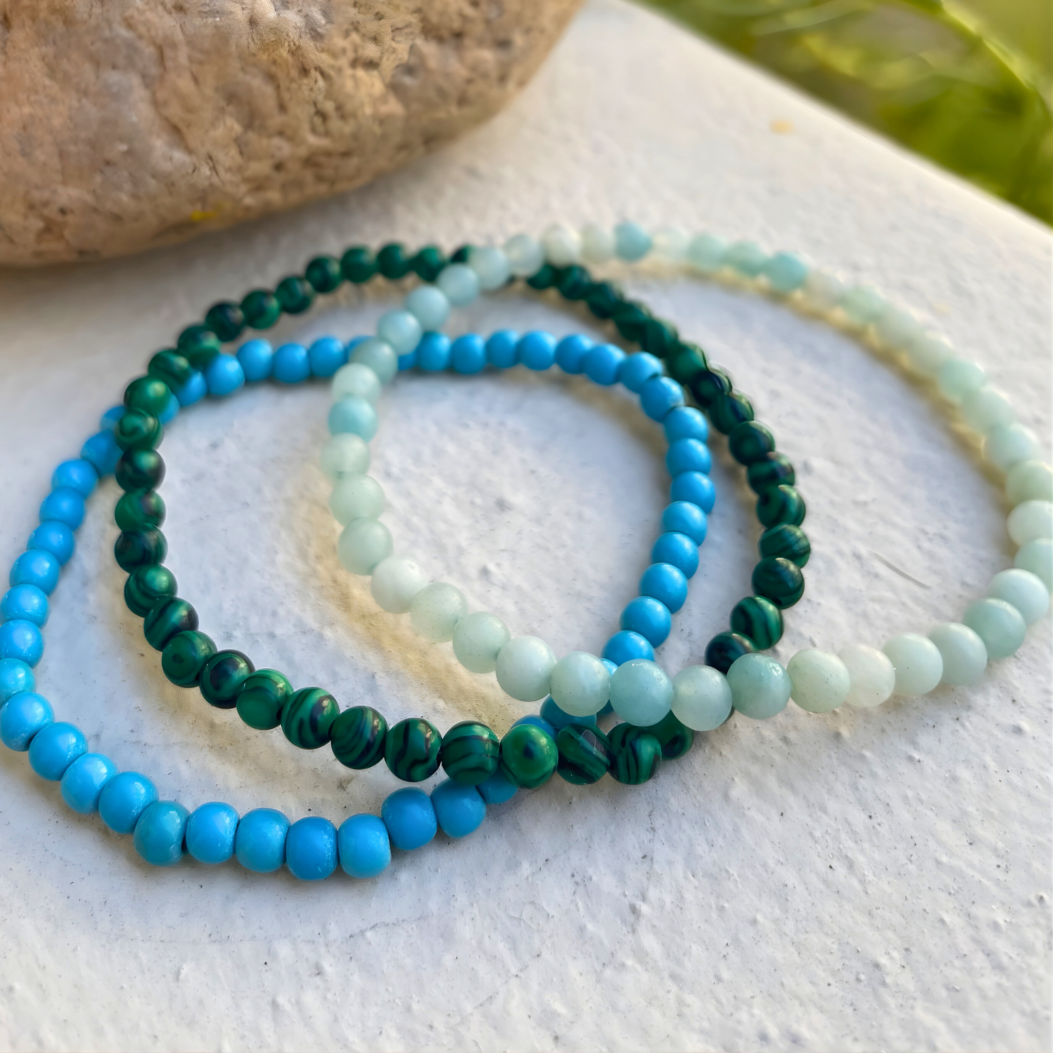 Three beaded bracelets in blue, green, and white on a textured surface.