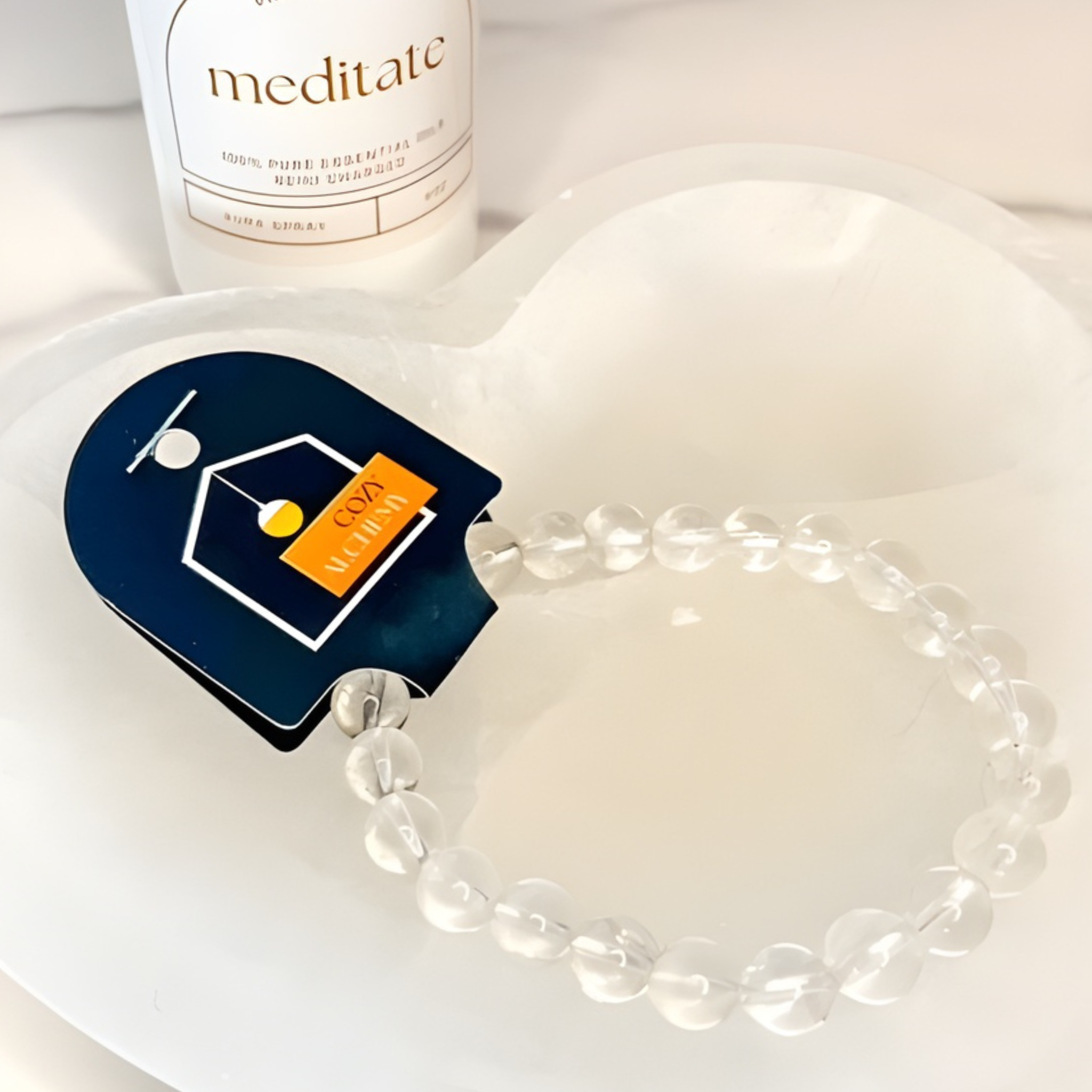White beaded bracelet on a white heart-shaped dish with a bottle labeled 'meditate' in the background.