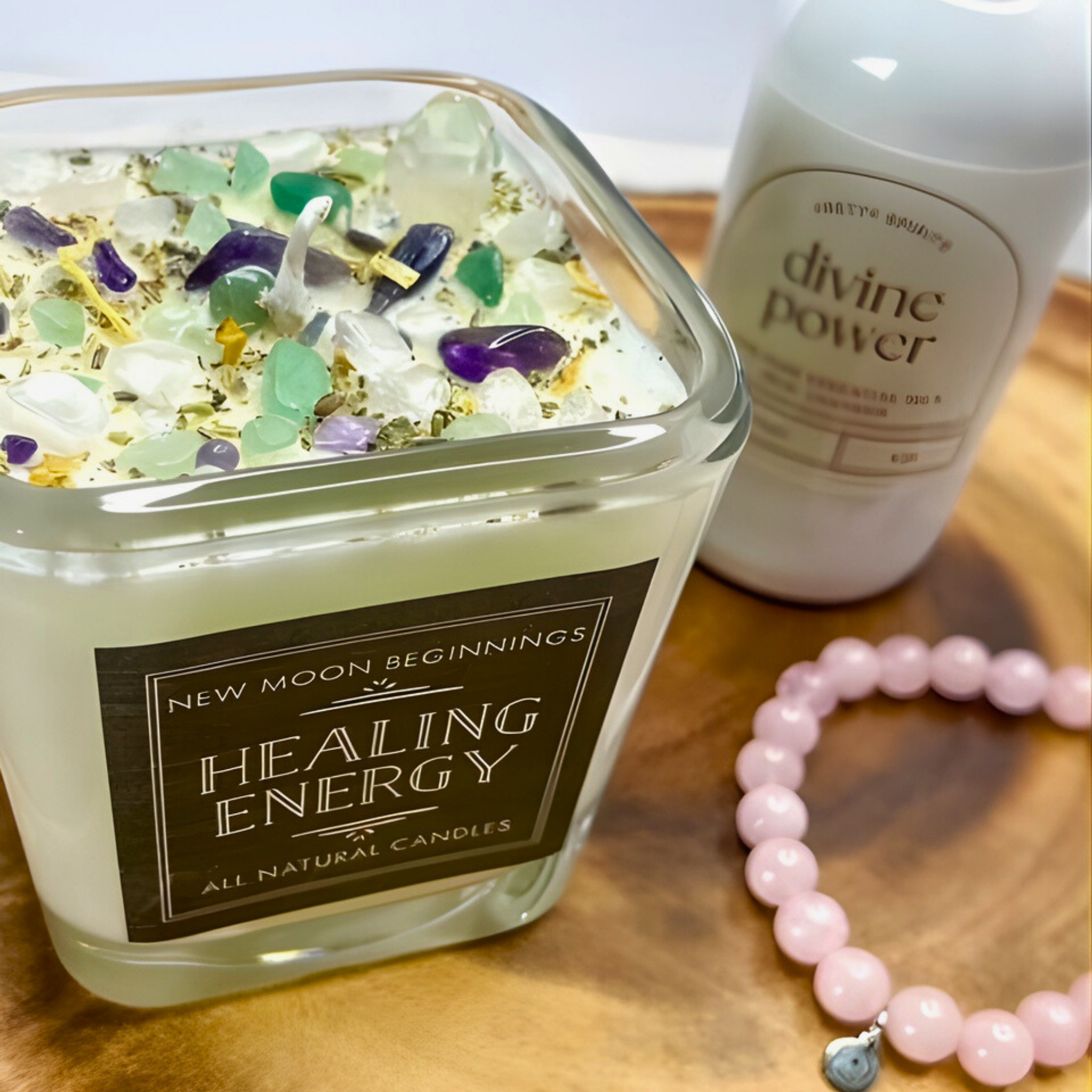 Healing energy candle with gemstones, bottle labeled 'divine power', and pink beaded bracelet on a wooden surface.