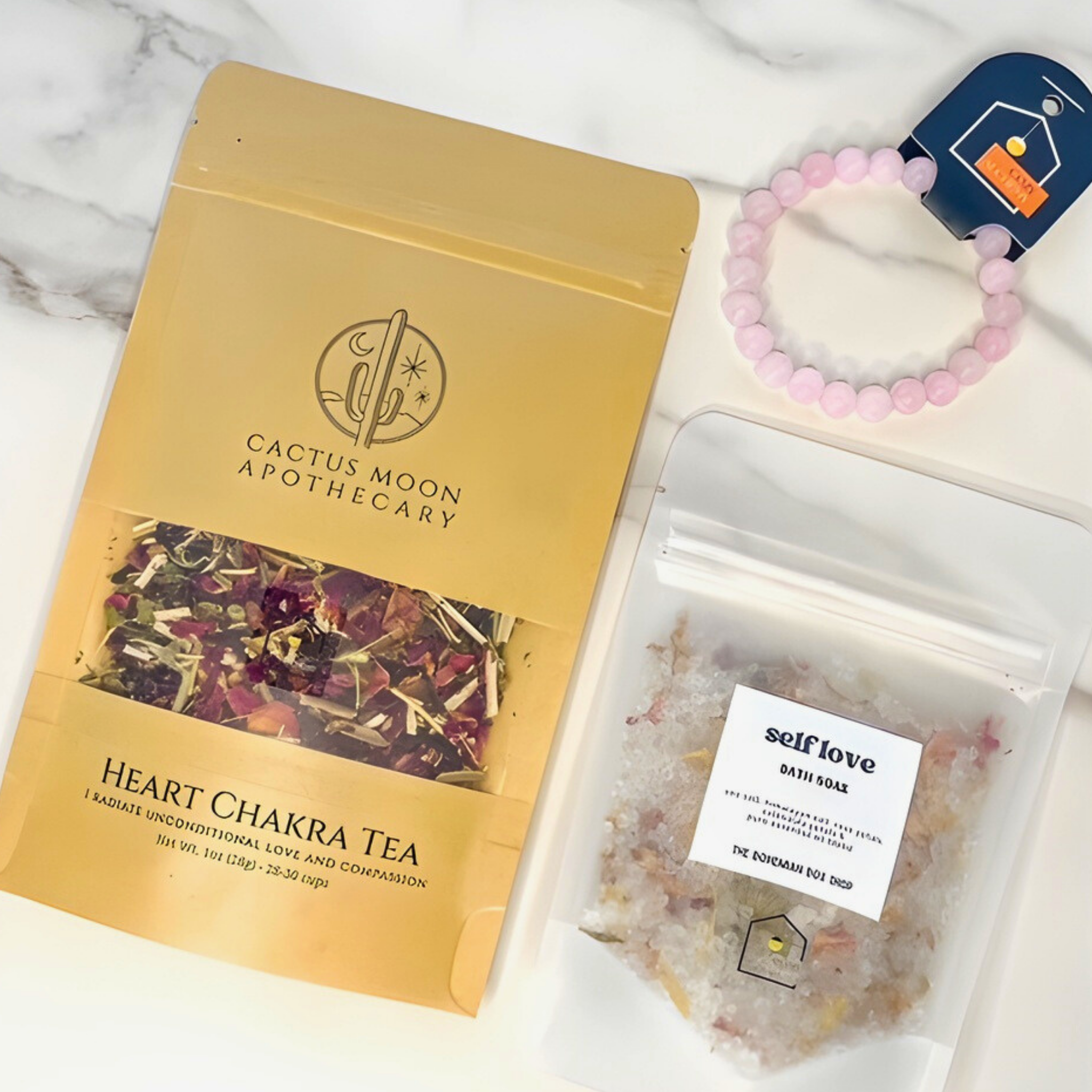 Cactus Moon Apothecary heart chakra tea package with a pink beaded bracelet and self-love bath soaks on a marble surface.