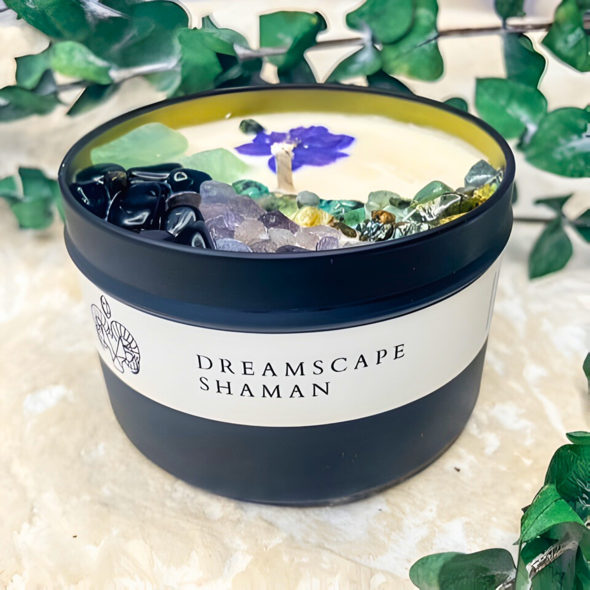 Candle in a container with decorative stones and a visible brand label on a natural background.