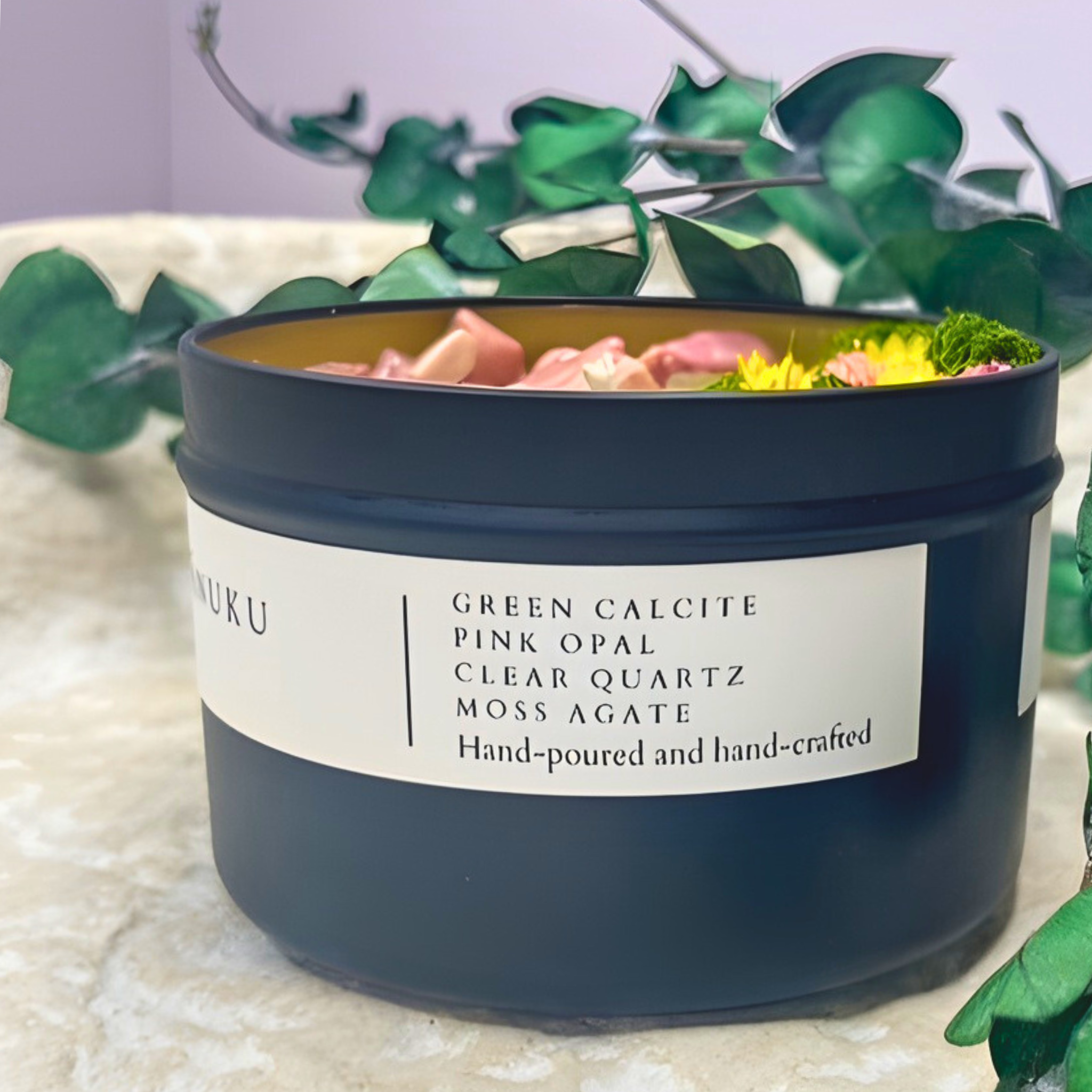 Candle in a black container with a label on a light surface with green leaves in the background