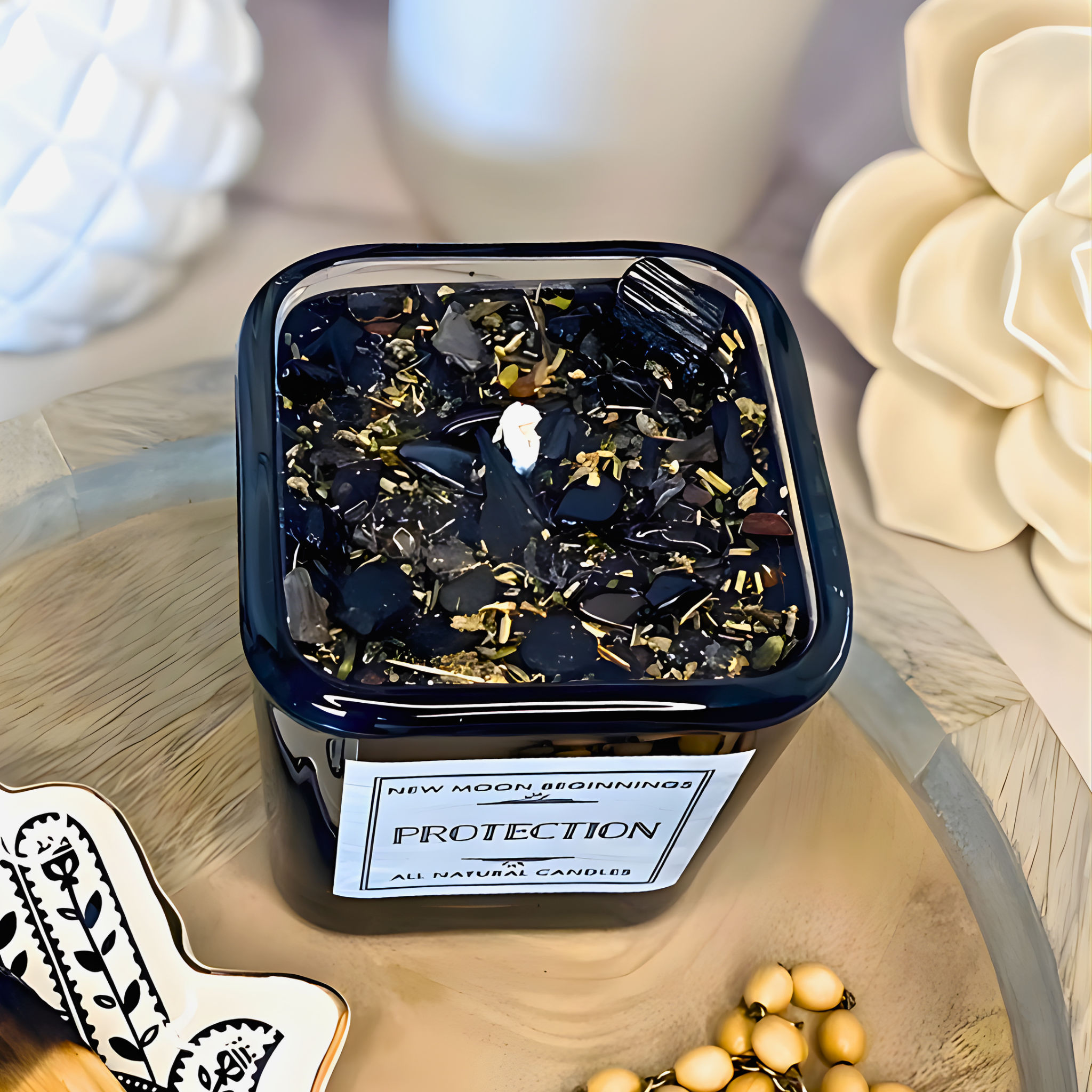 Container of New Moon Beginnings Protection candle on a decorative surface with flowers and jewelry.