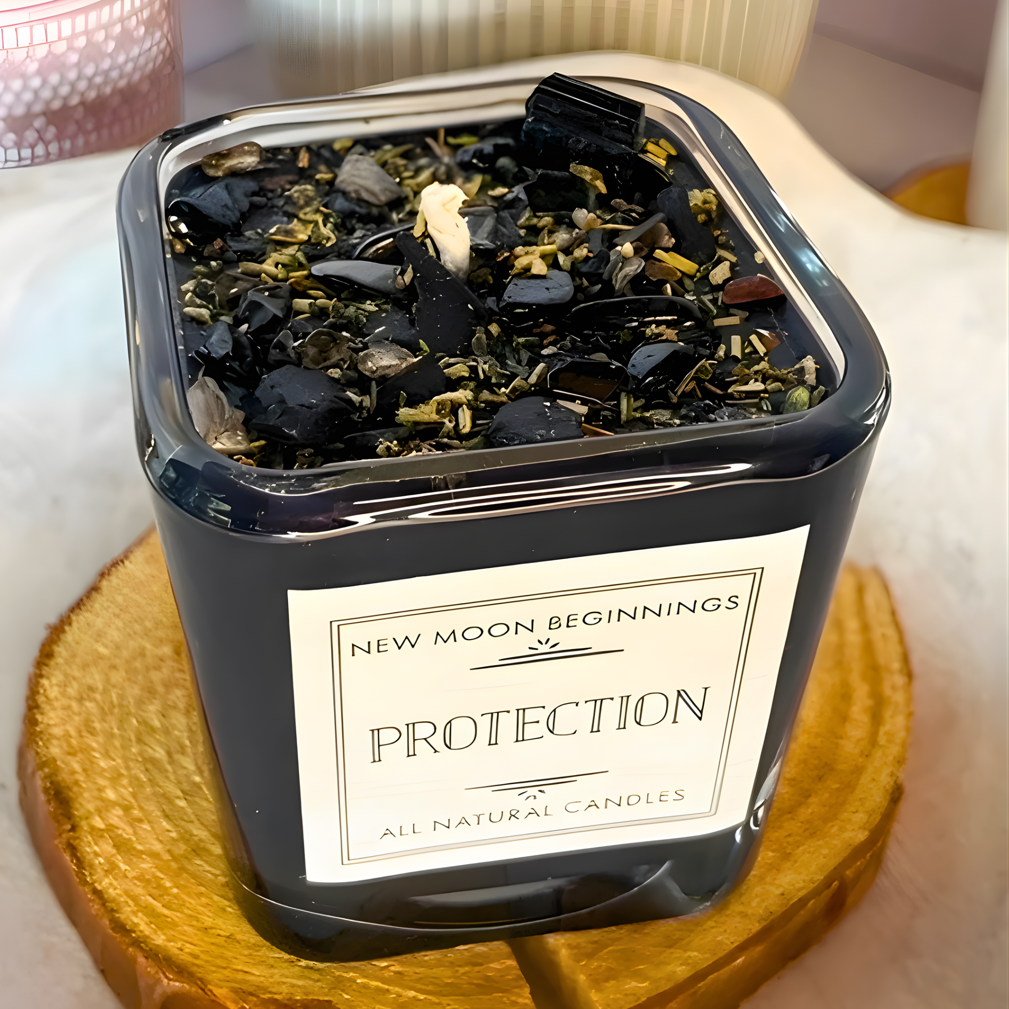 Candle in a black container with decorative elements on a wooden coaster, labeled 'New Moon Beginnings Protection'.