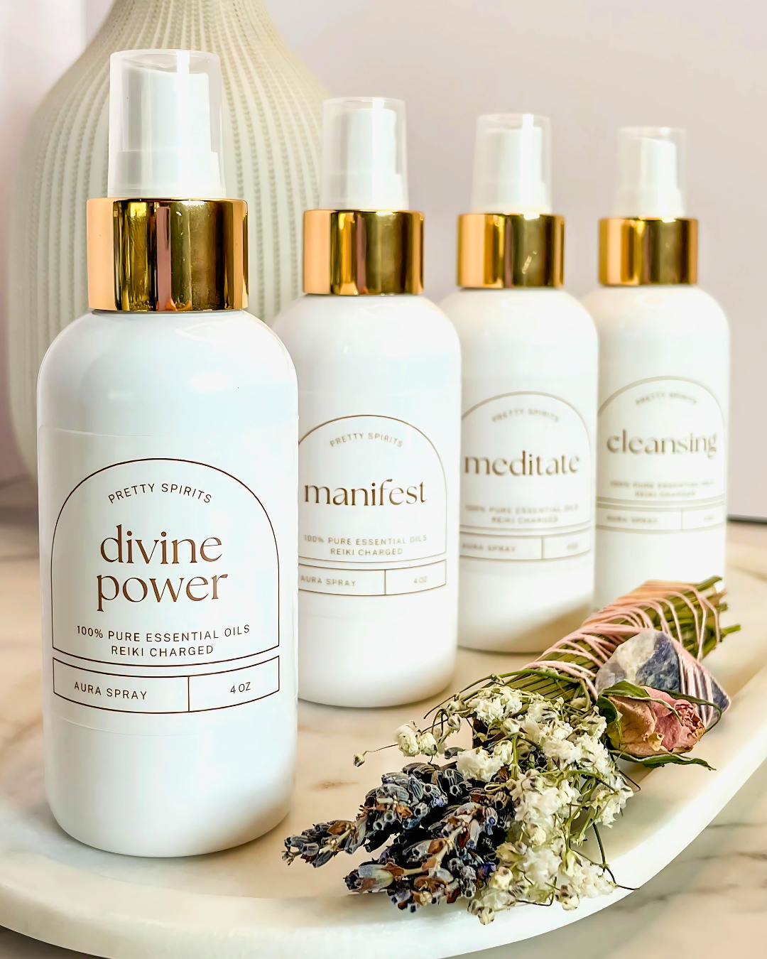Four white bottles with gold caps labeled 'divine power', 'manifest', 'meditate', and 'cleansing' on a marble surface with dried flowers.
