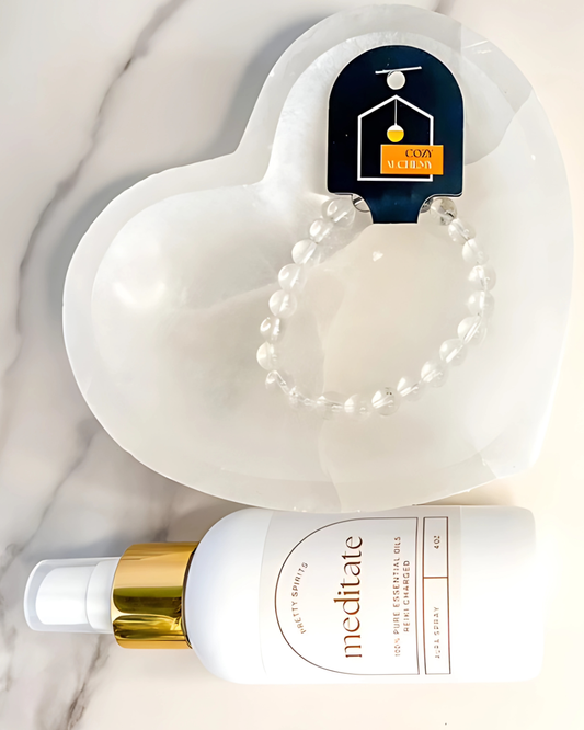 White bottle with gold cap labeled 'meditate' on a marble surface with a heart-shaped object and bracelet in the background.