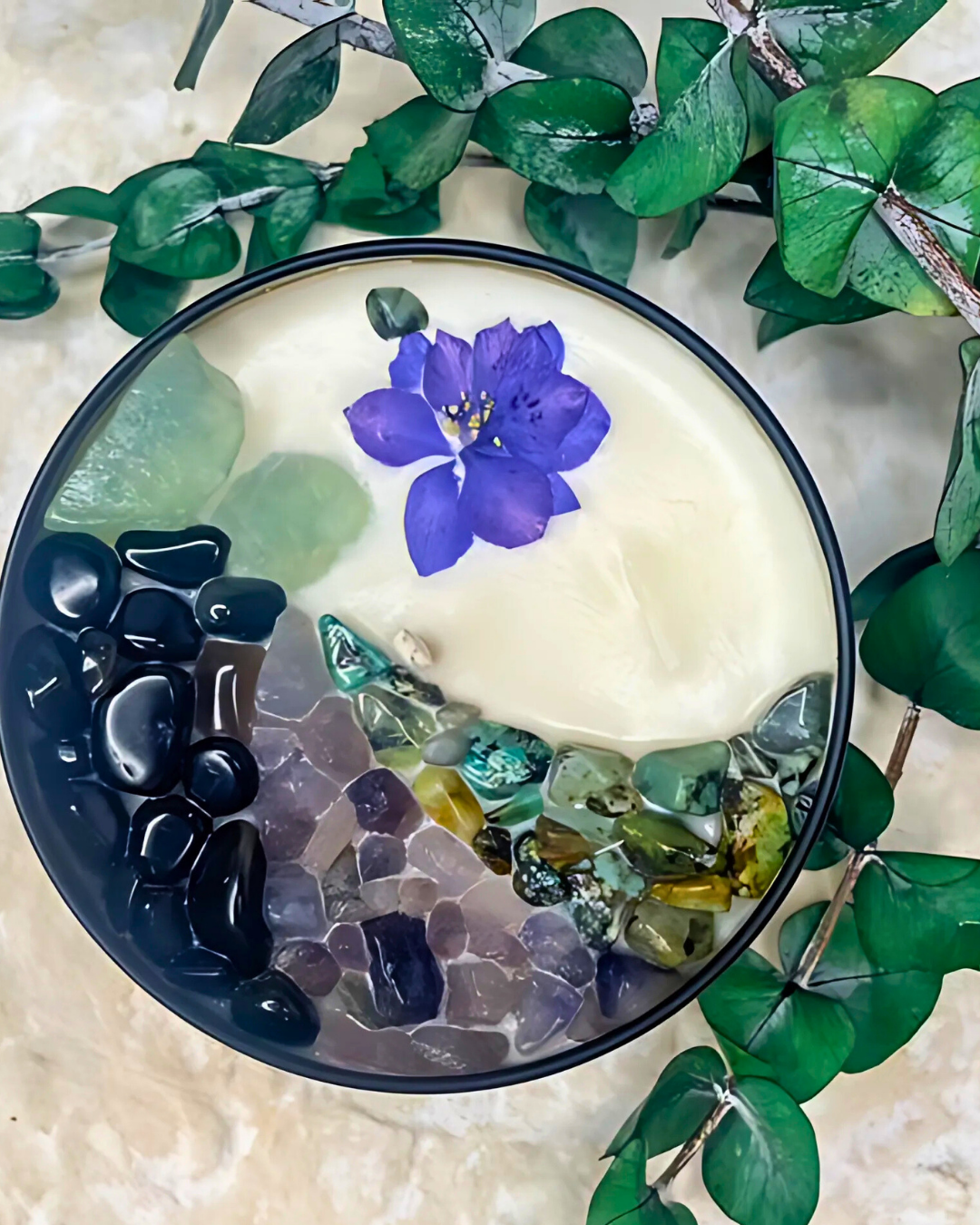 Decorative dish with stones, a purple flower, and green leaves on a textured surface.