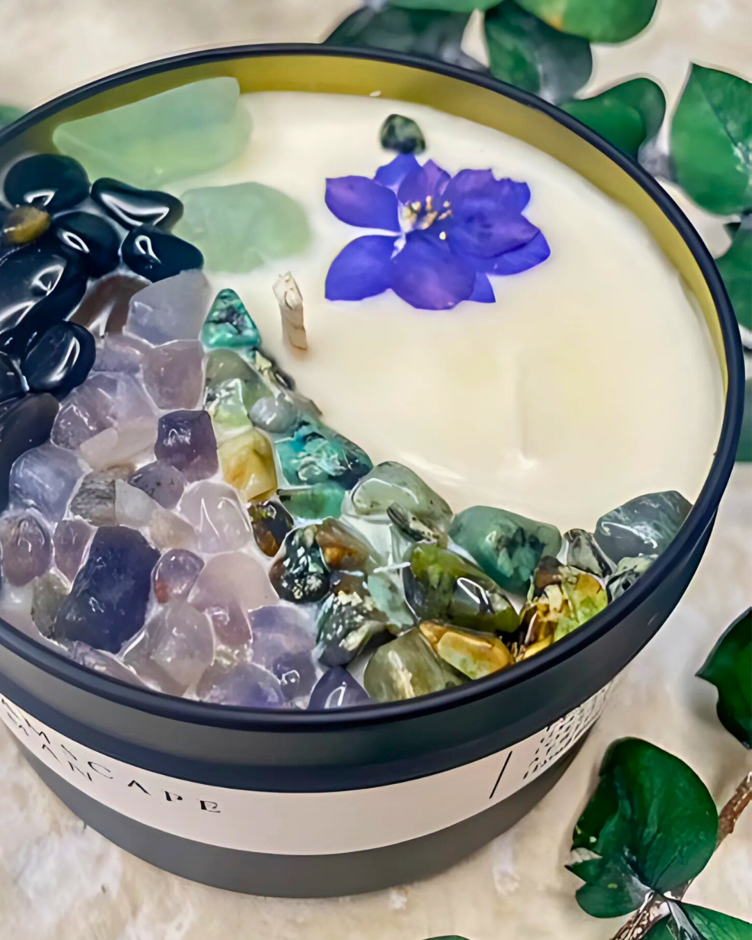 Candle with embedded stones and a purple flower, surrounded by green leaves.