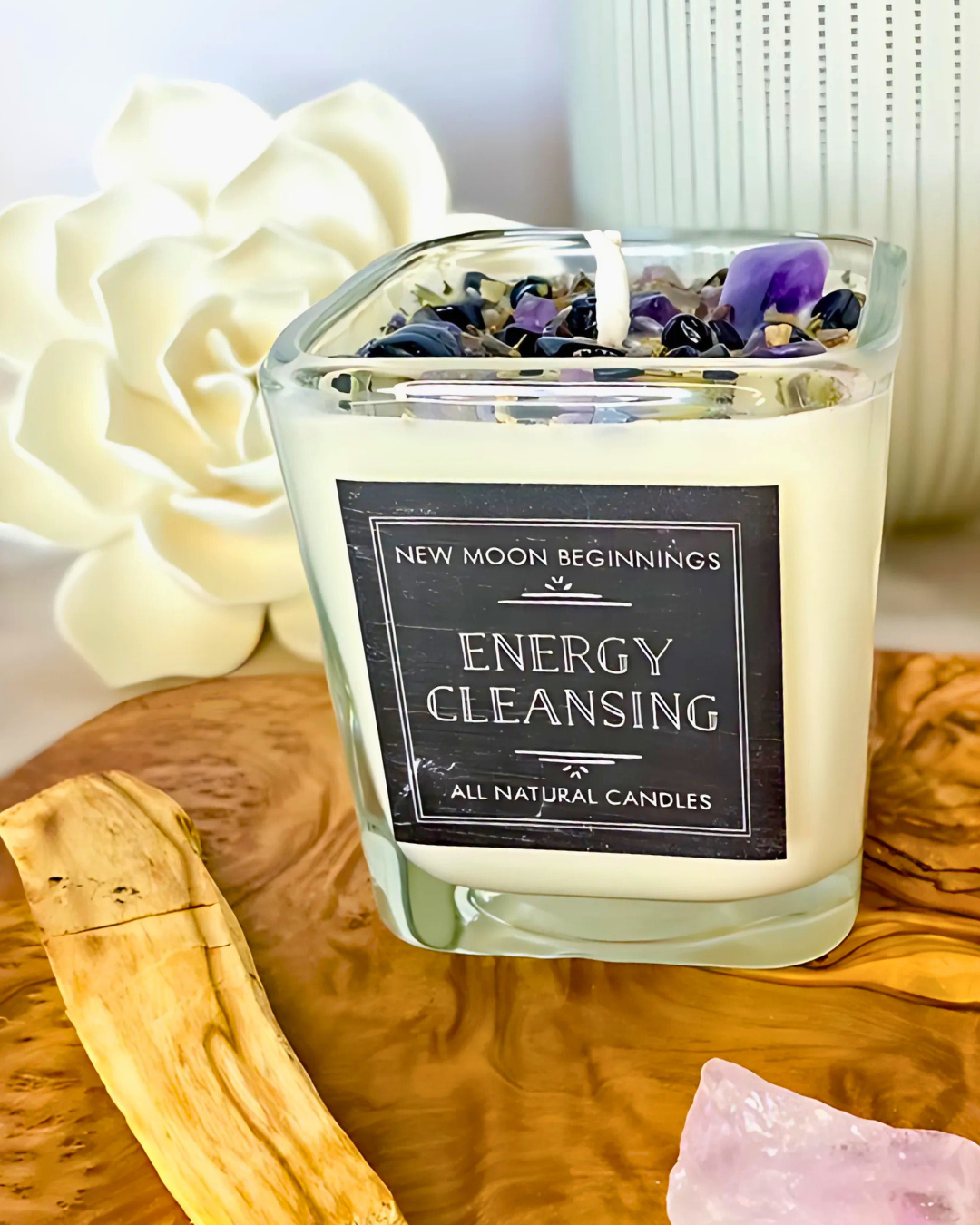 Candle labeled 'Energy Cleansing' from 'New Moon Beginnings' on a wooden surface with flowers and crystals.