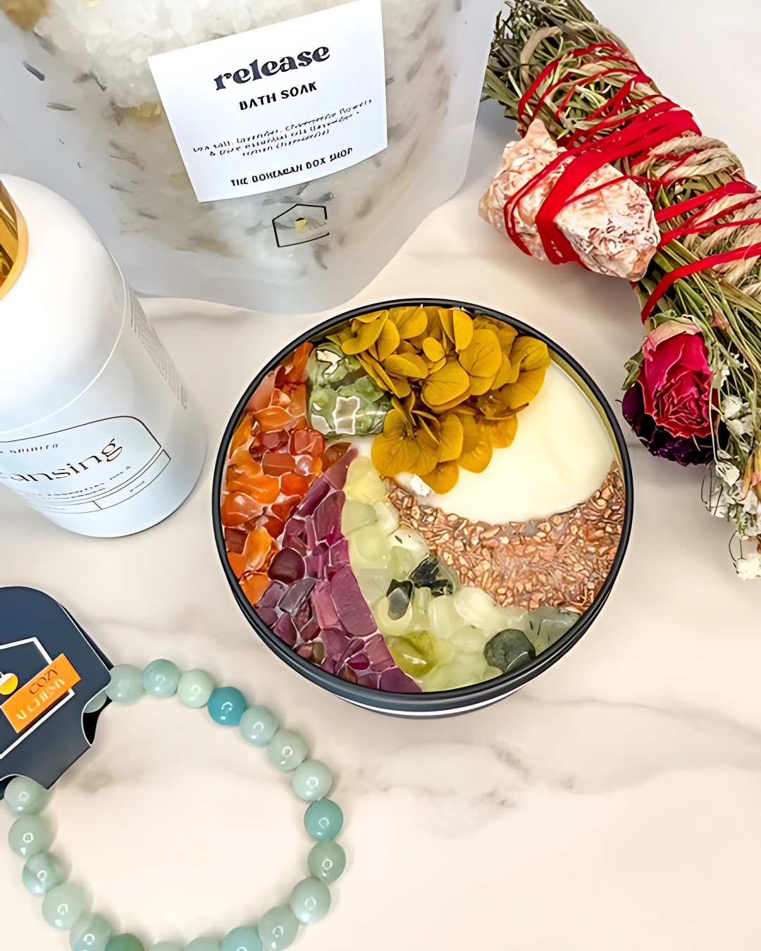 Colorful bath bomb bowl with a candle, 'release' bath soap, and smudge stick on a marble surface.