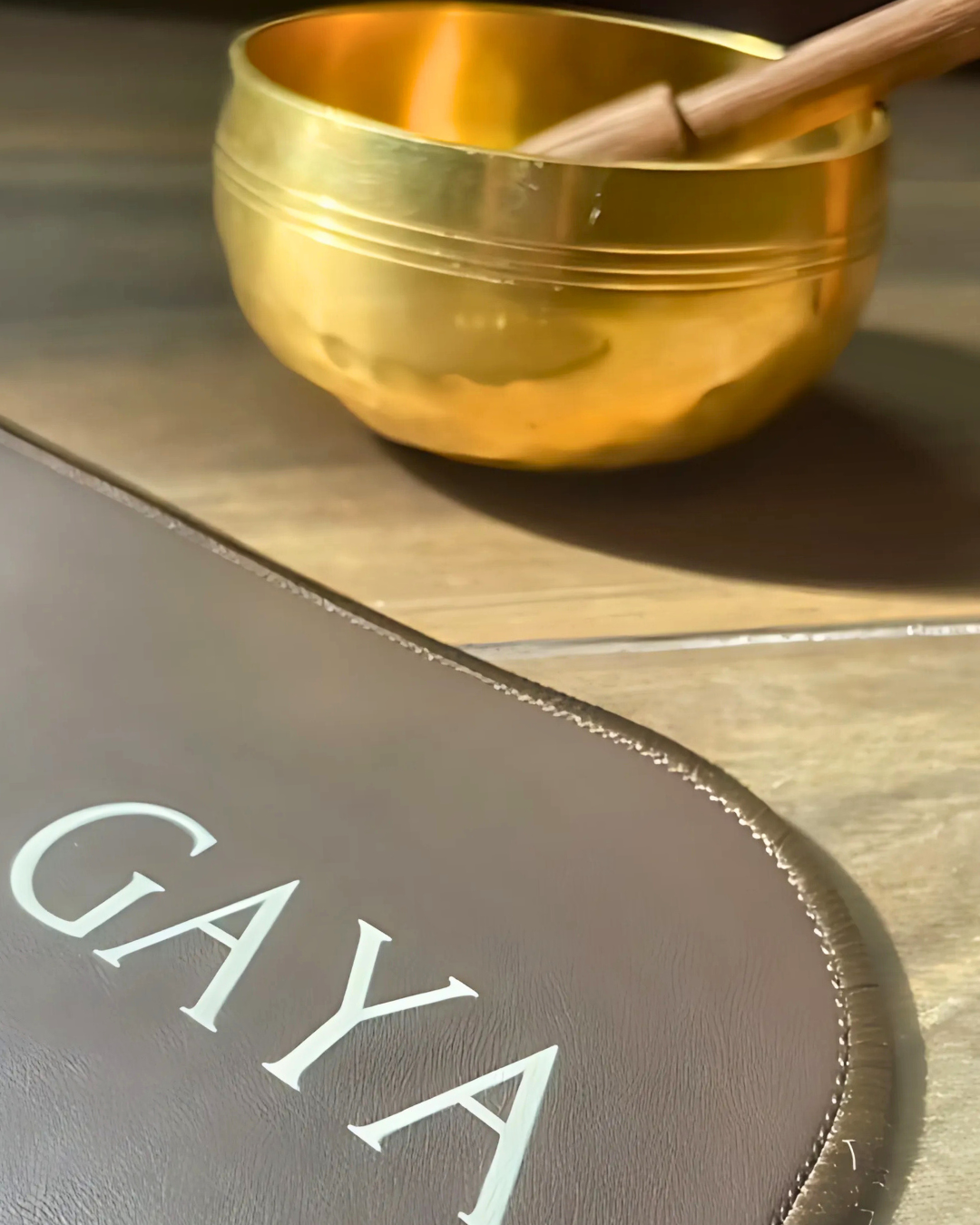 Gold singing bowl with a wooden stick on a wooden surface, with 'GAYA' branding visible.