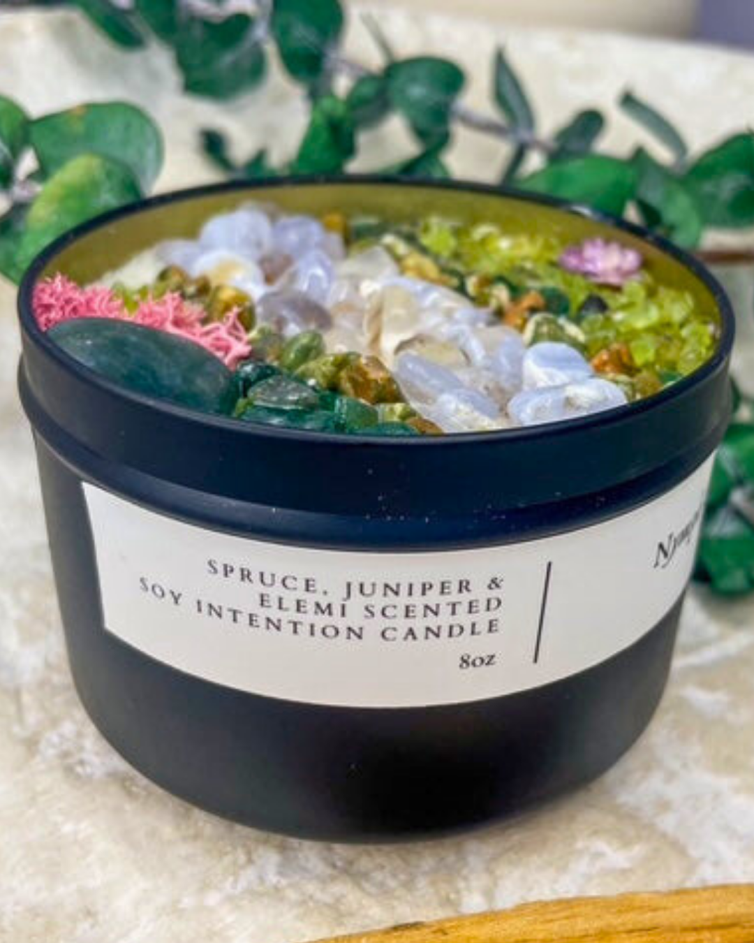 Scented candle with decorative stones and herbs in a black container on a white background.