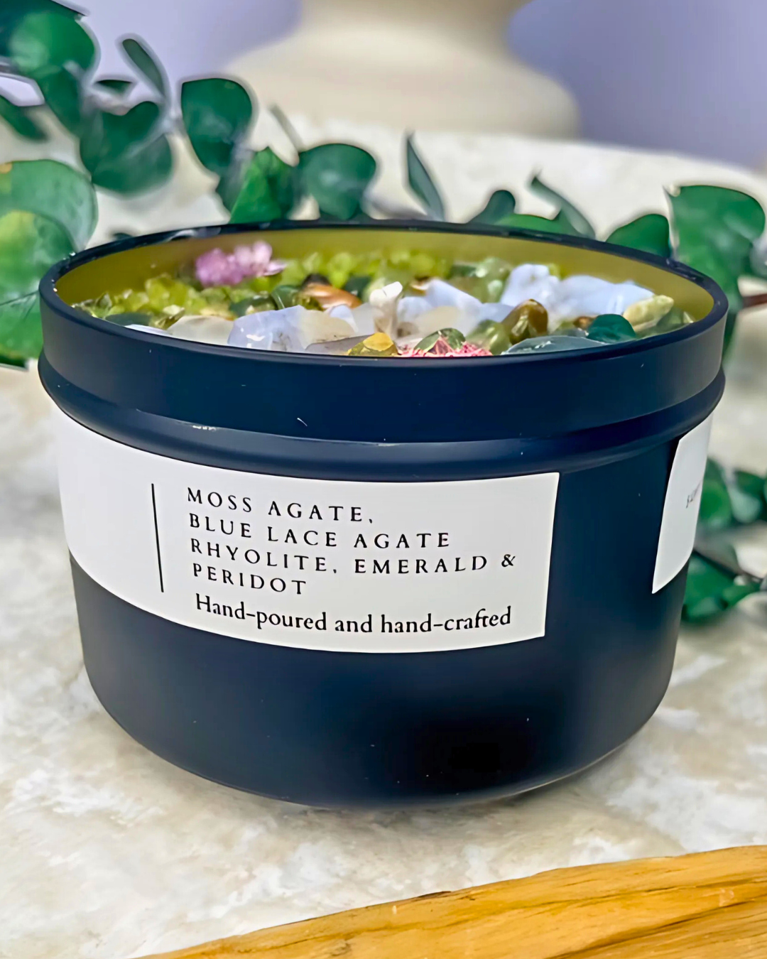 Candle in a black container with decorative stones on a textured surface with greenery in the background