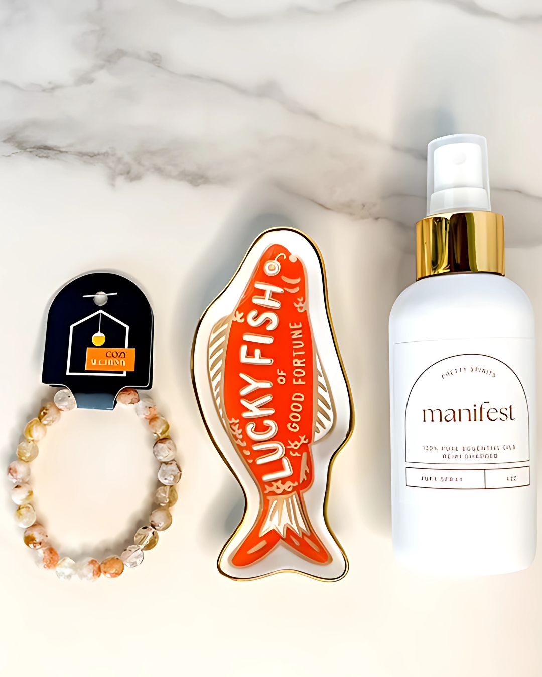 Bracelet, 'Lucky Fish' ornament, and 'manifest' spray bottle on a marble surface