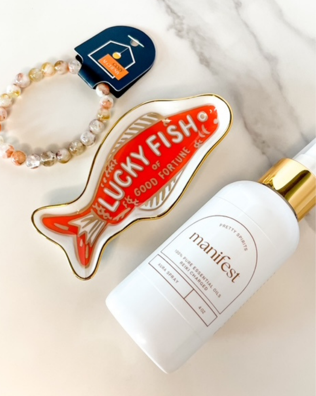 Lucky Fish Good Fortune pin next to a bottle labeled 'manifest' on a marble surface.