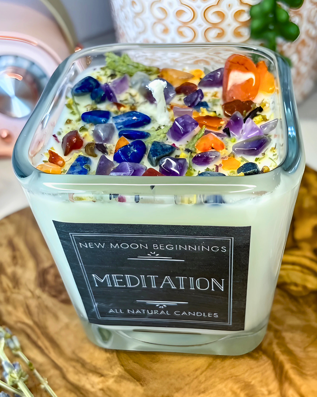 Candle with colorful stones and 'New Moon Beginnings Meditation' label on a wooden surface