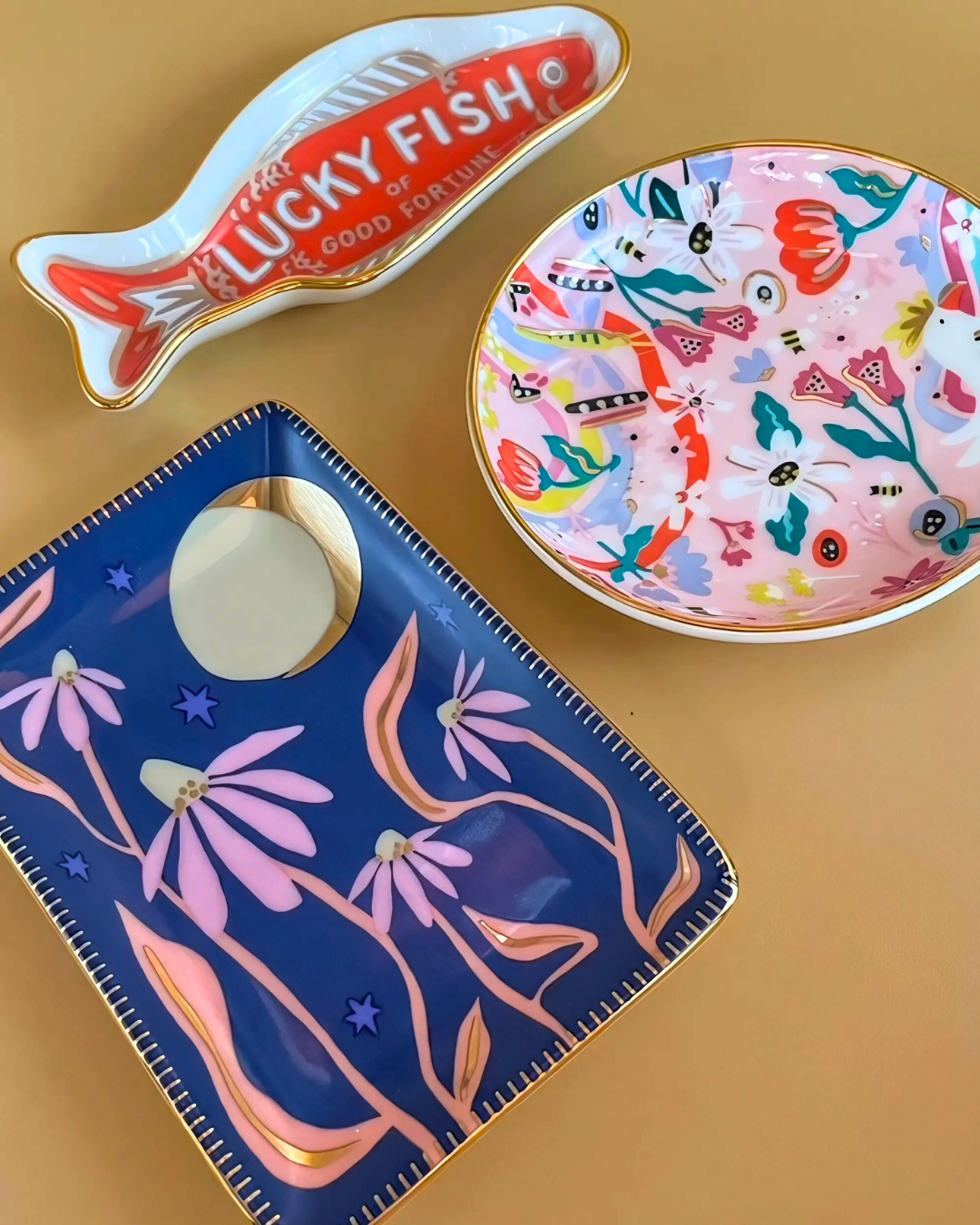 Three decorative items on a beige surface: a fish-shaped tray, a colorful plate with floral design, and a blue pouch with pink floral patterns.