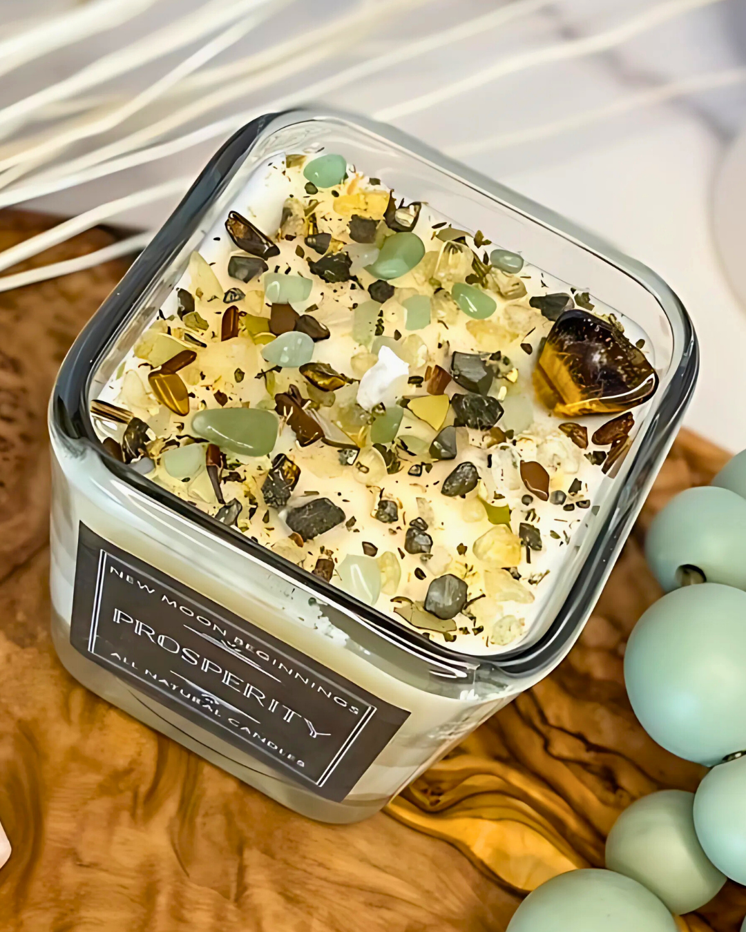 Candle with decorative stones in a clear container on a wooden surface with beads.