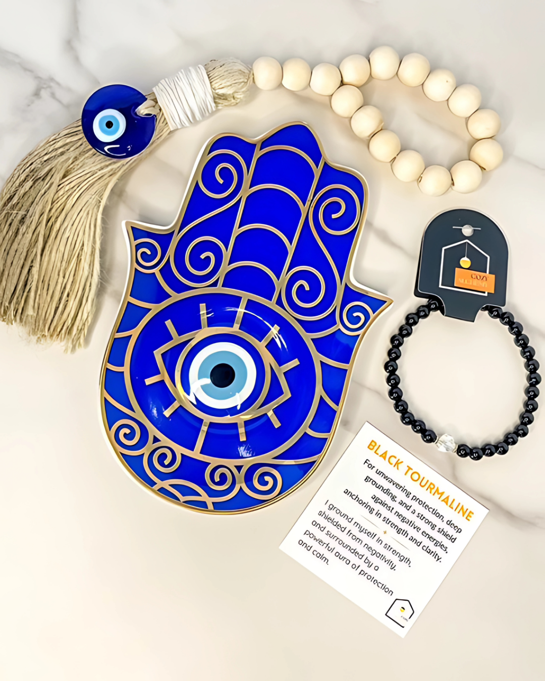 Blue Hamsa hand with evil eye, tassel, and black tourmaline bracelet on a marble surface.
