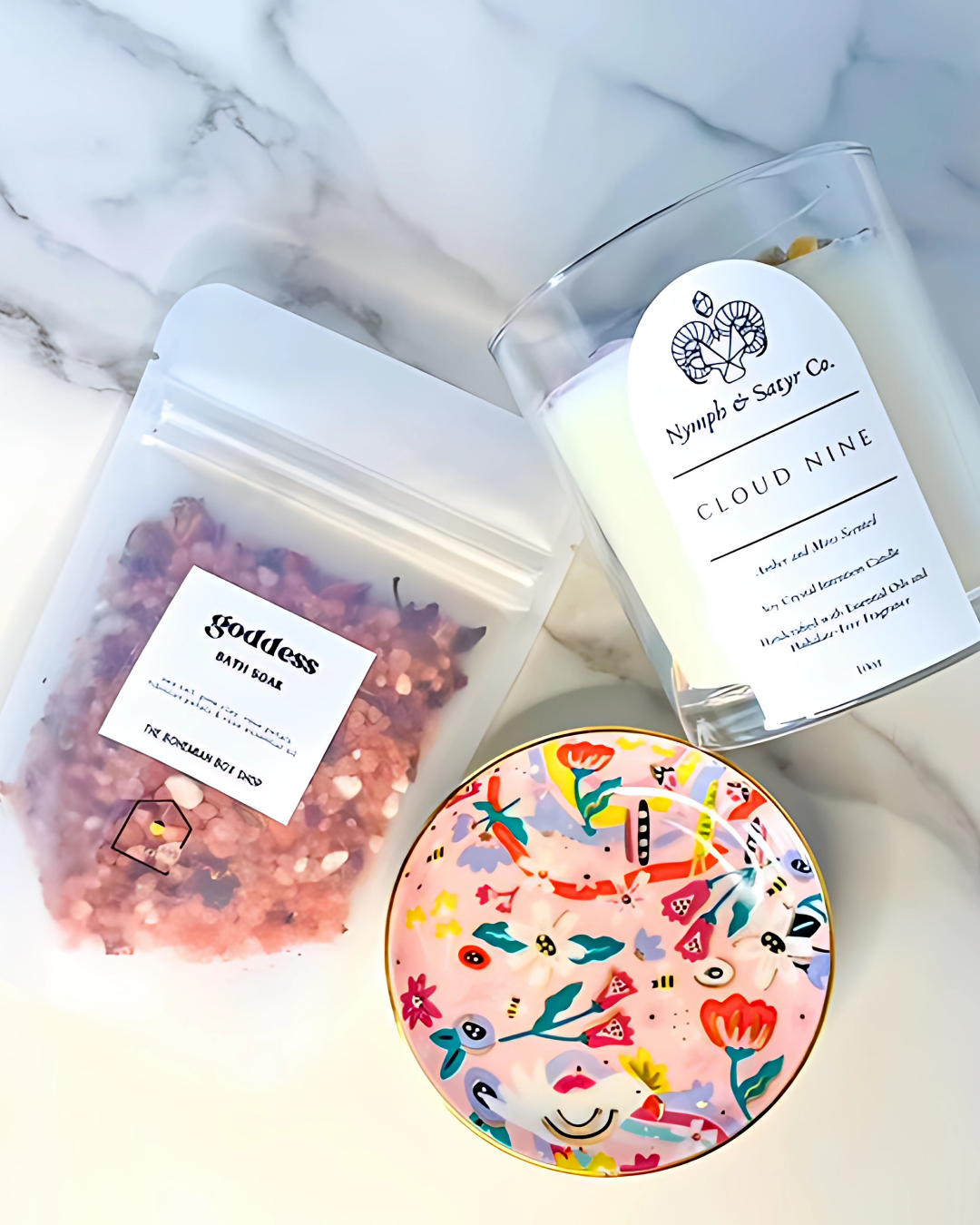 Candle labeled 'Cloud Nine' next to a bag labeled 'Goddess' and a decorative round item with floral design on a marble surface.