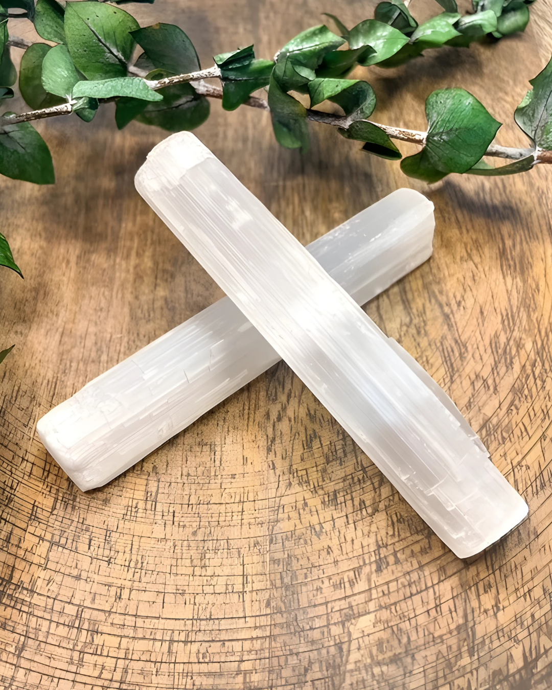 Two selenite sticks on a wooden surface with green leaves in the background
