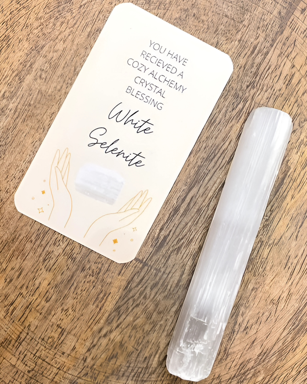 White selenite crystal on a wooden surface with a card reading 'Cozy Alchemy Crystal Blessing'.