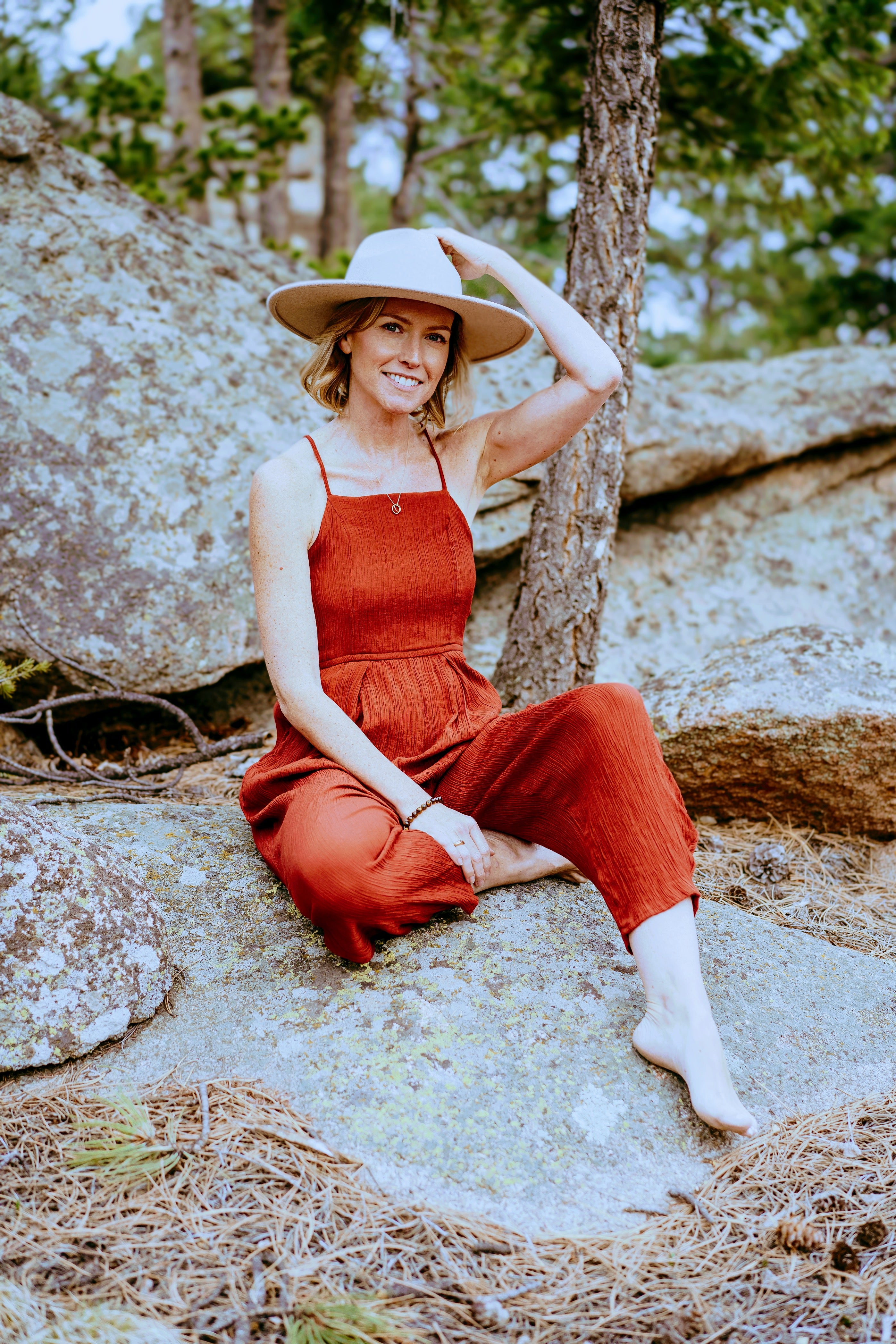 Woman in a red jumpsuit and white hat sitting on rocks with trees in the background