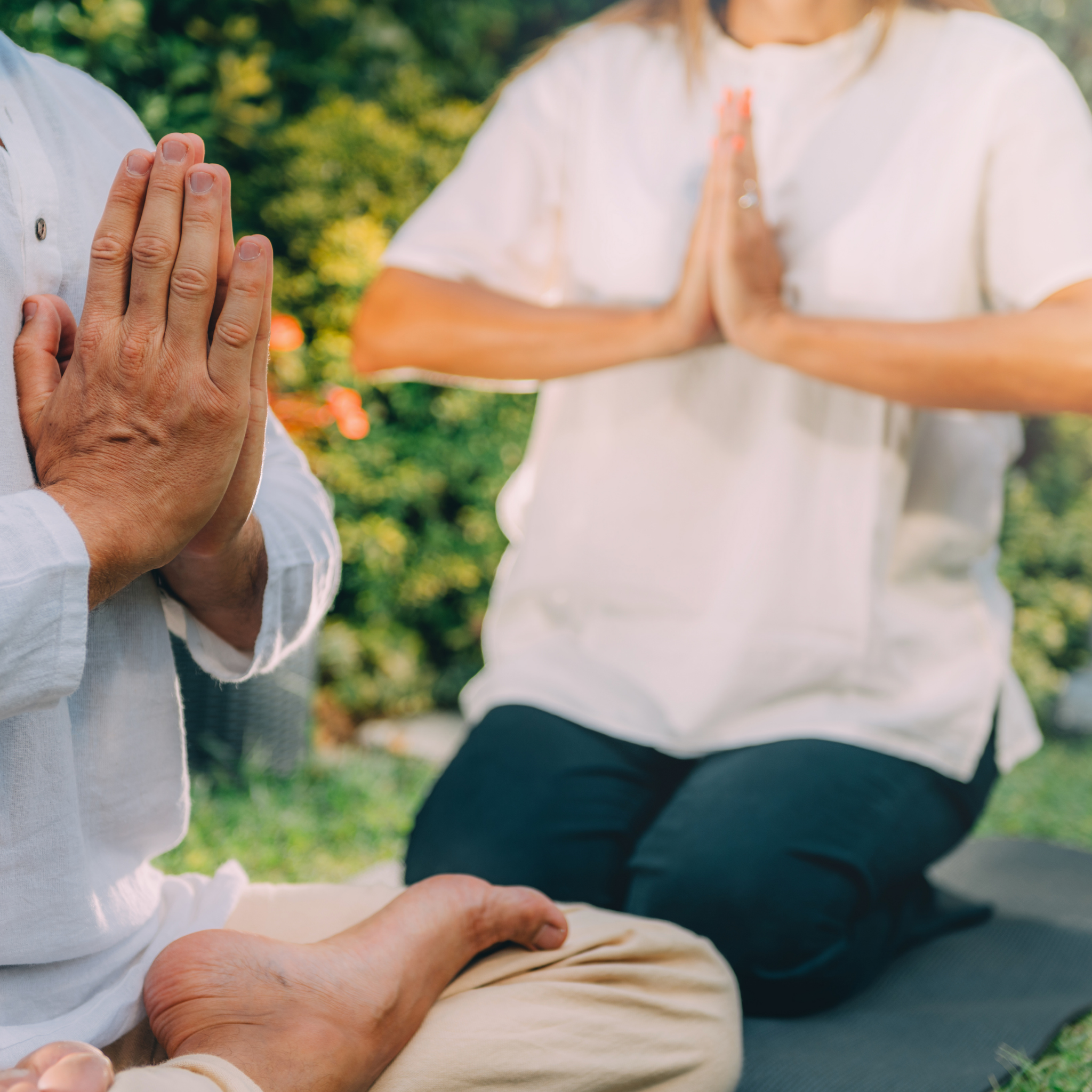 Two people sitting outdoors with hands pressed together, possibly in a meditative pose.